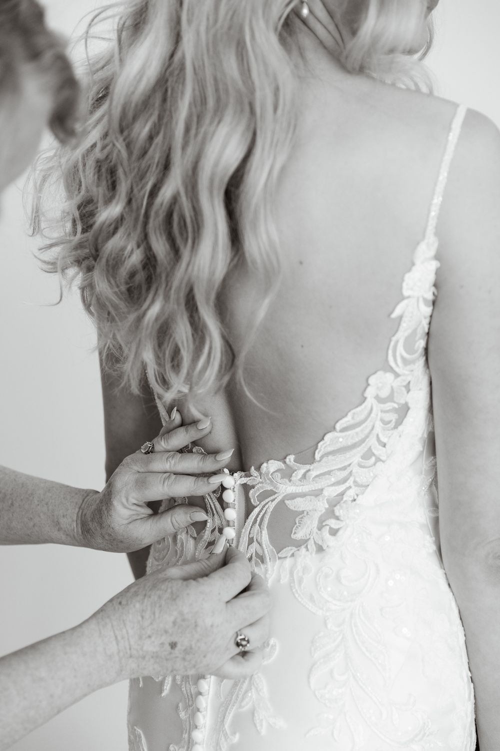 Black and white photo of bride in lace backless wedding gown being buttoned up by another person.