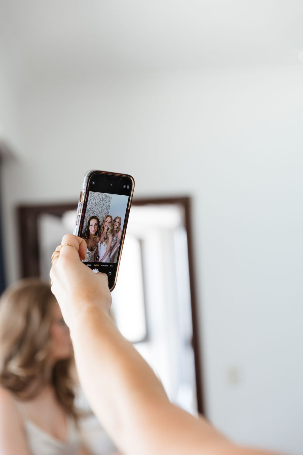 Woman holding smartphone taking a group selfie with bridesmaids reflected in mirror during wedding preparations.