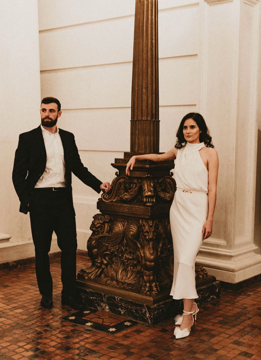 A couple poses formally beside an ornate carved pillar base in an elegant hallway.