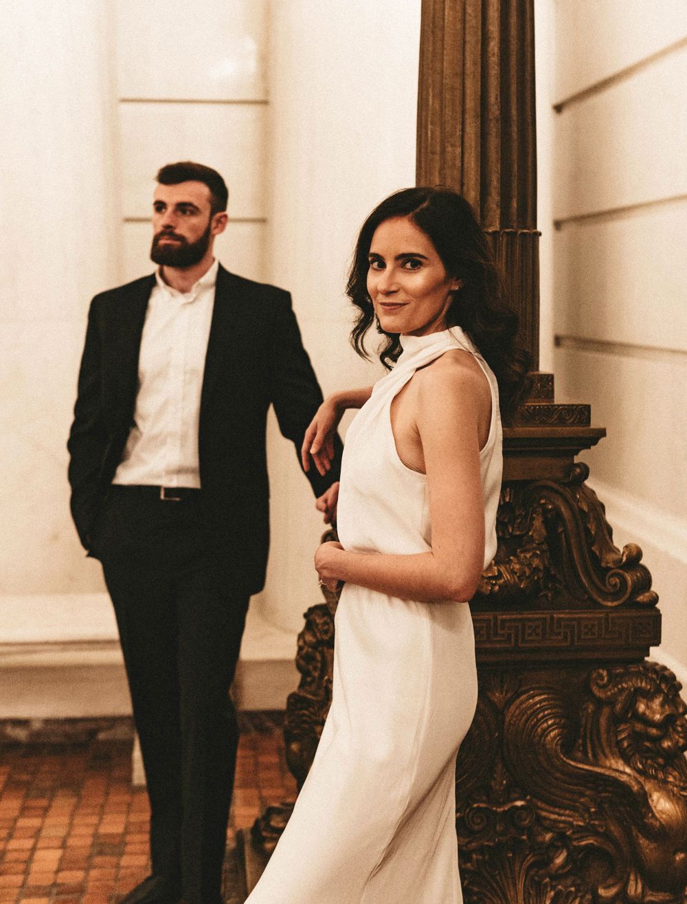 A stylish couple poses beside an ornately carved column in formal attire.