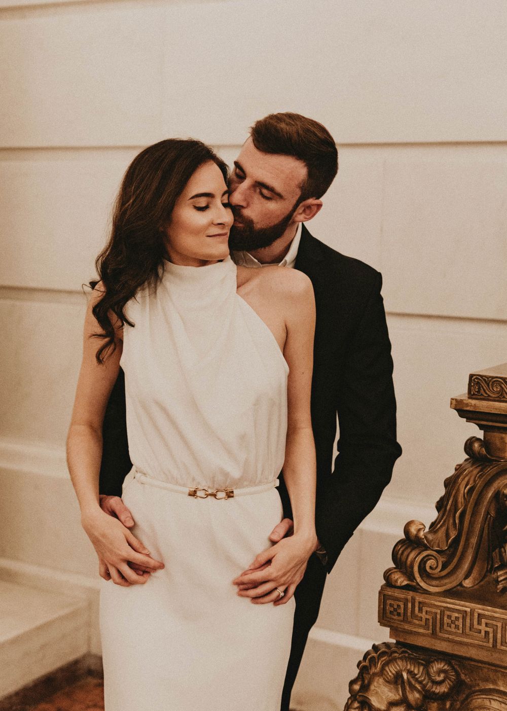 An intimate portrait of a couple in formal wear against a light-colored wall.