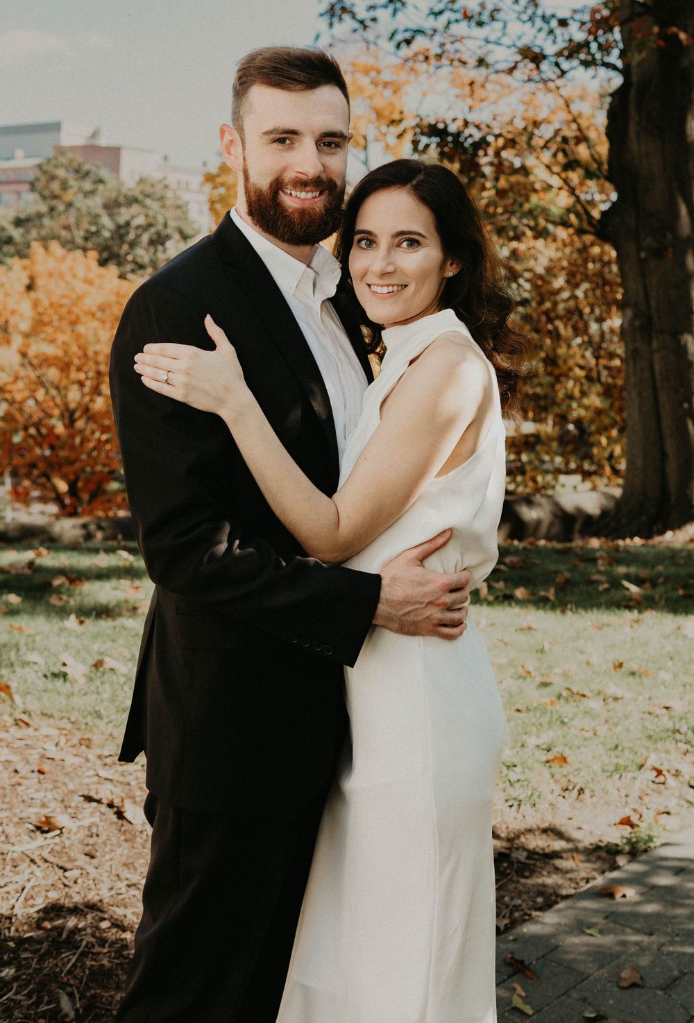 A couple poses outdoors for their wedding photos during autumn with fall foliage in the background.
