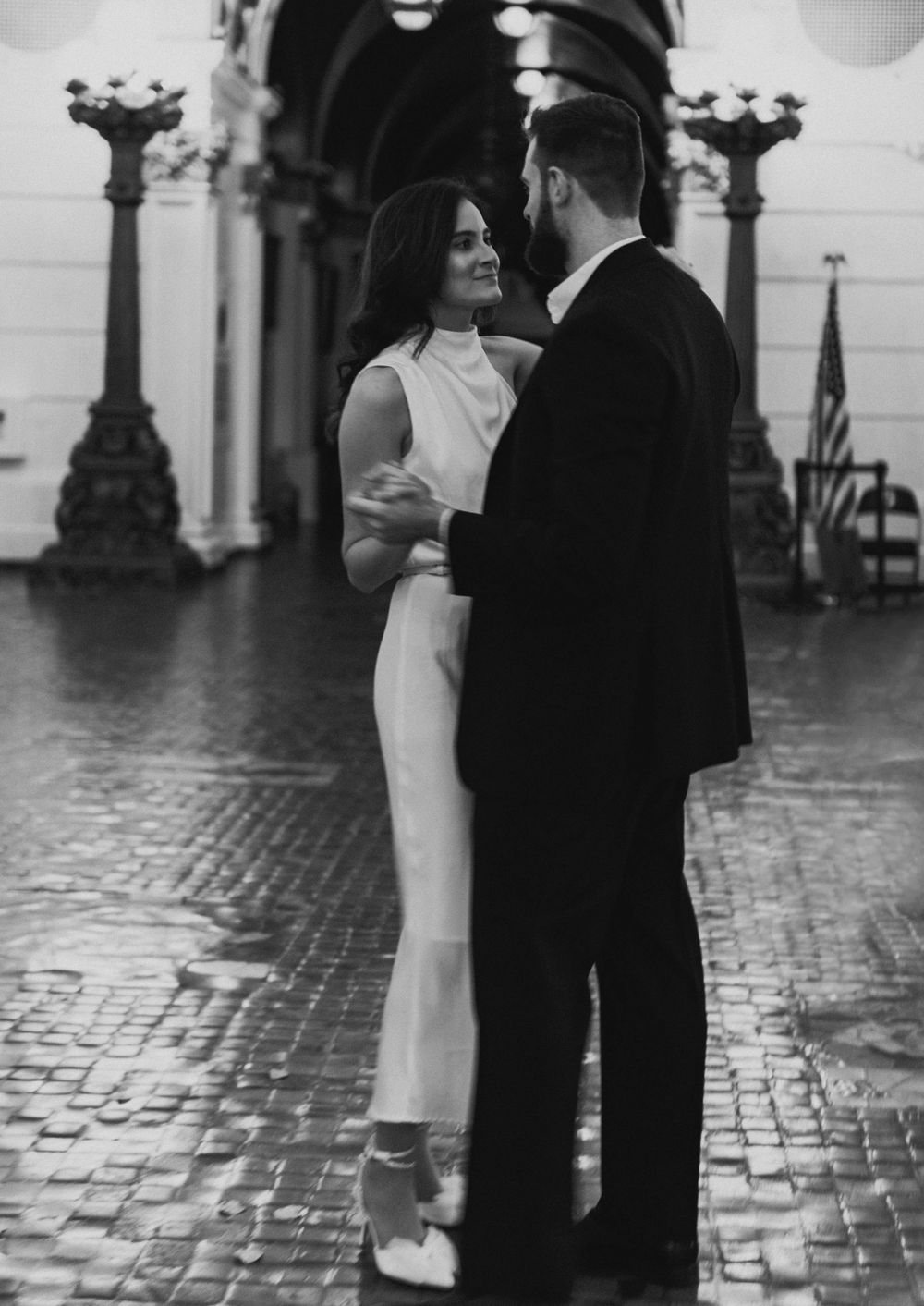 A black and white photo of a couple in formal attire standing on ornate cobblestones.