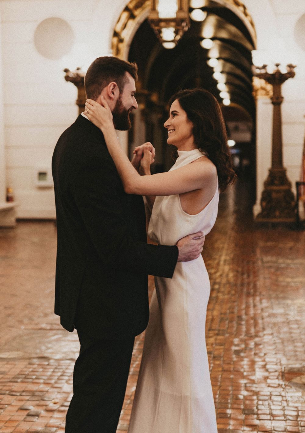 A romantic couple embraces in an elegant hallway with ornate architecture and warm lighting.