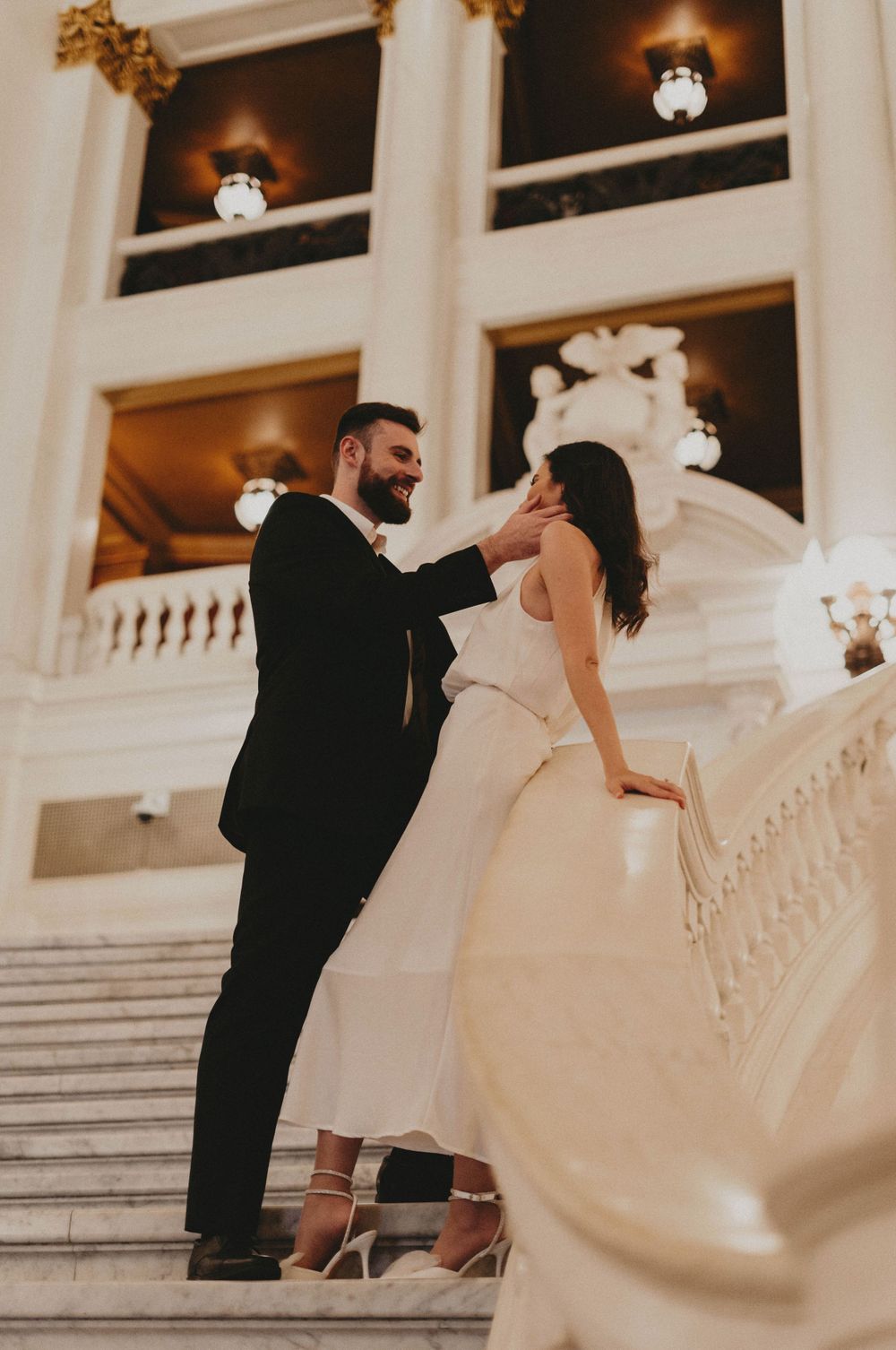 A romantic moment captured on a grand white marble staircase with ornate lighting fixtures.