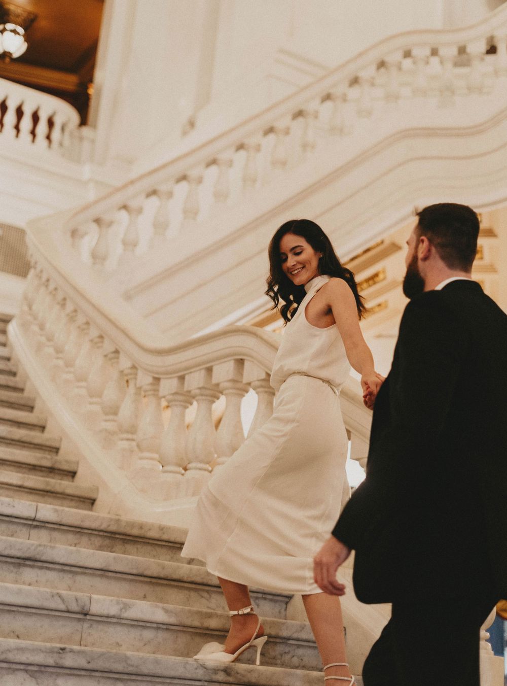 A couple ascends an elegant marble staircase in formal attire.