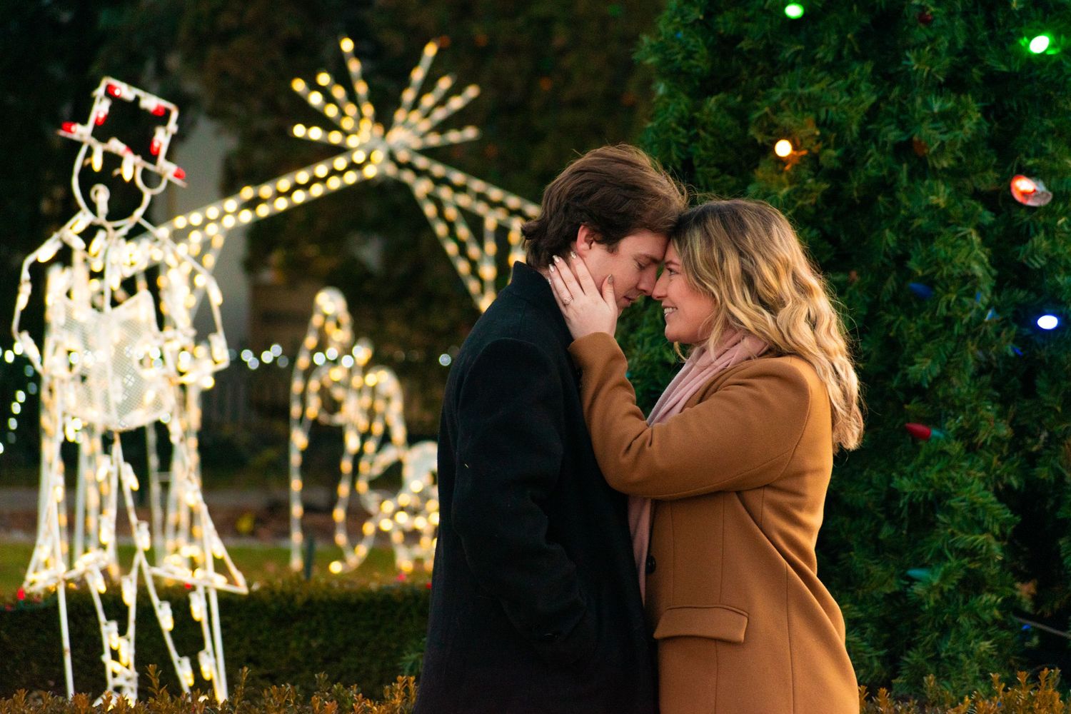 A tender embrace captured against a backdrop of holiday lights and illuminated reindeer decorations.