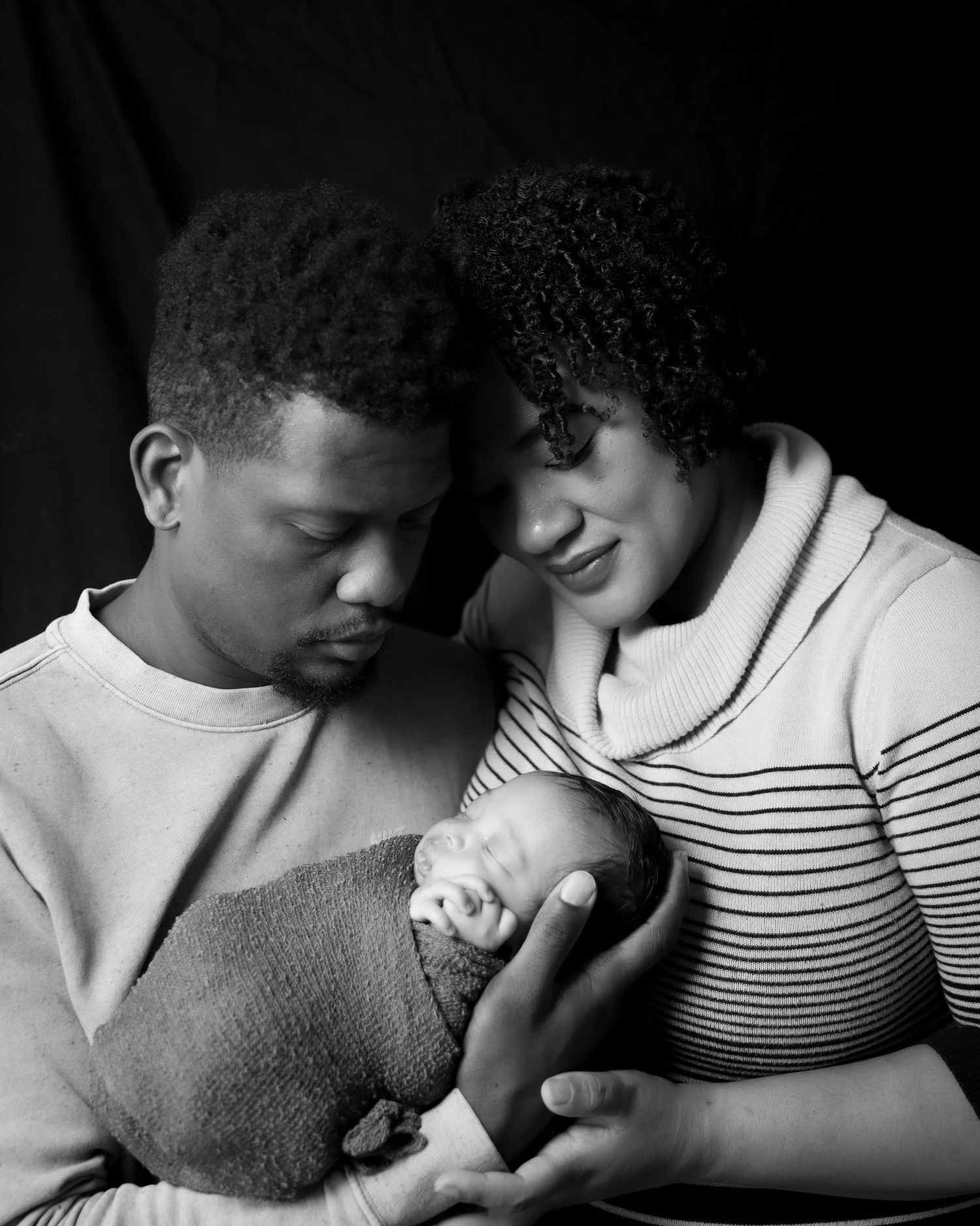 Intimate black and white portrait of couple cradling newborn against dark background.