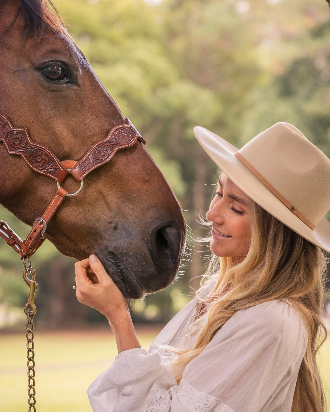 A brown horse and person in a white dress and tan hat share a tender moment in golden sunlight.