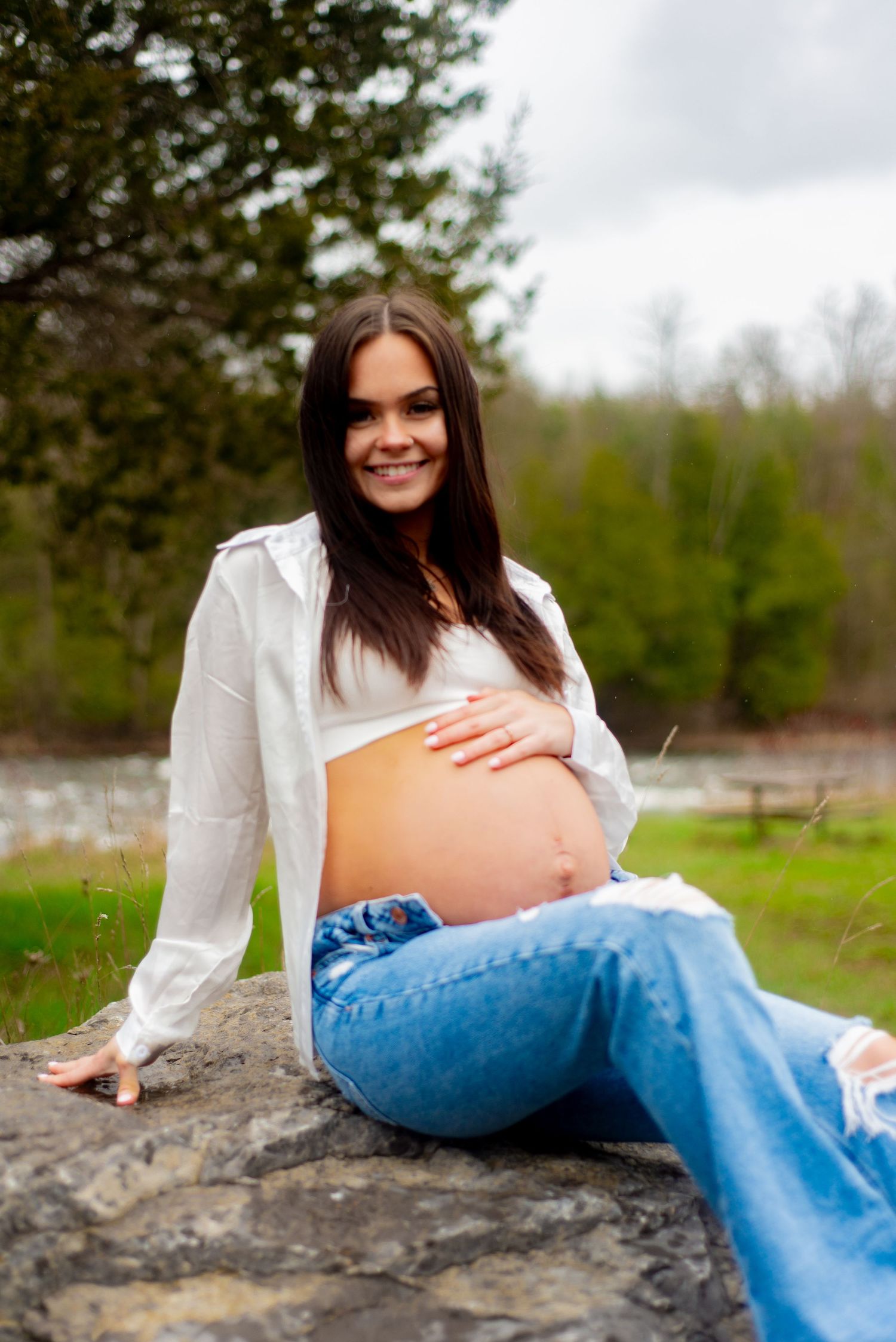Natural maternity portrait showing woman in white shirt on rock with forest background.