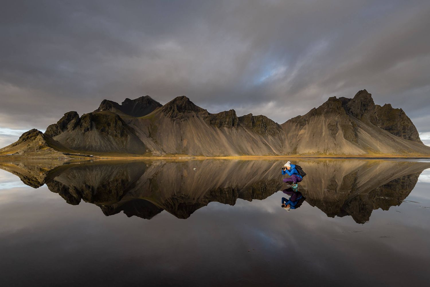 Photographer crouching beside perfect mountain reflection in still Icelandic lake under dramatic sky.