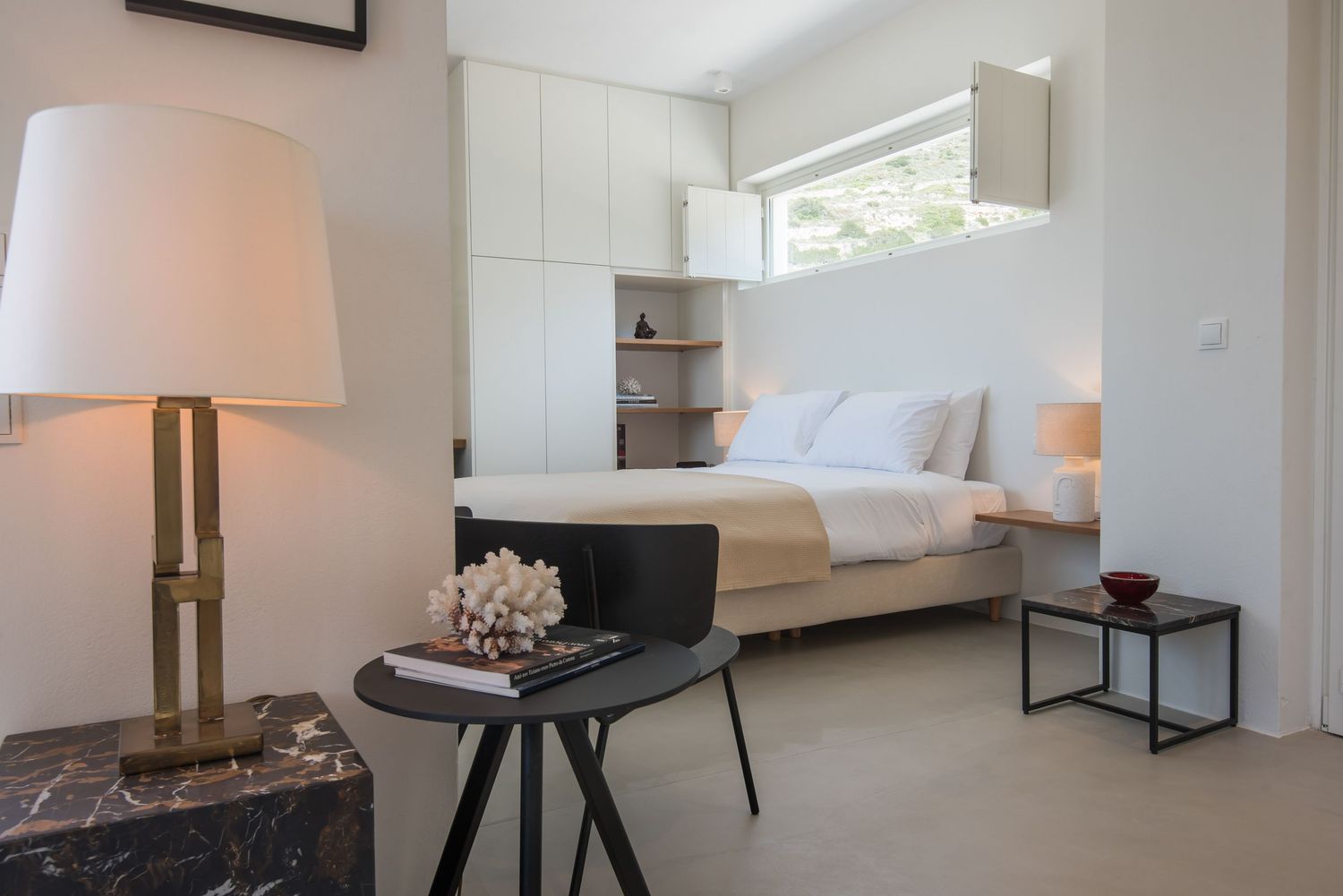 Modern bedroom interior with white bedding, tall windows, and a decorative table lamp on dark marble side table. Paros suites
