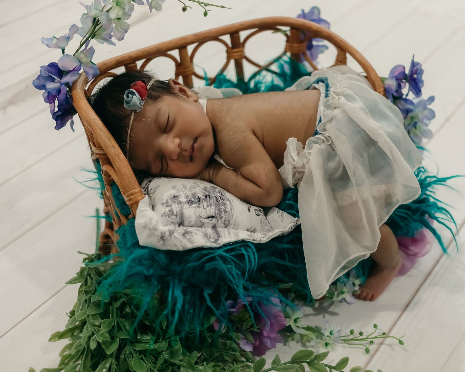 Sleeping newborn in a wicker basket decorated with teal fabric and purple flowers.