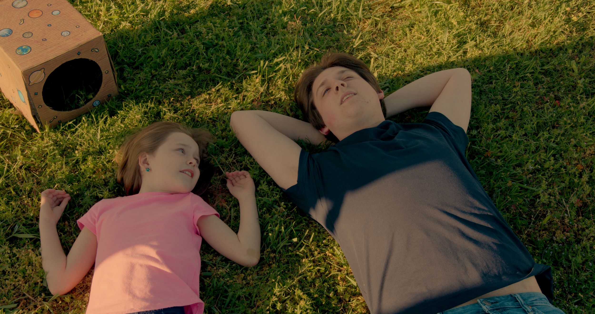 Two people lying in green grass next to a cardboard box on a sunny day.