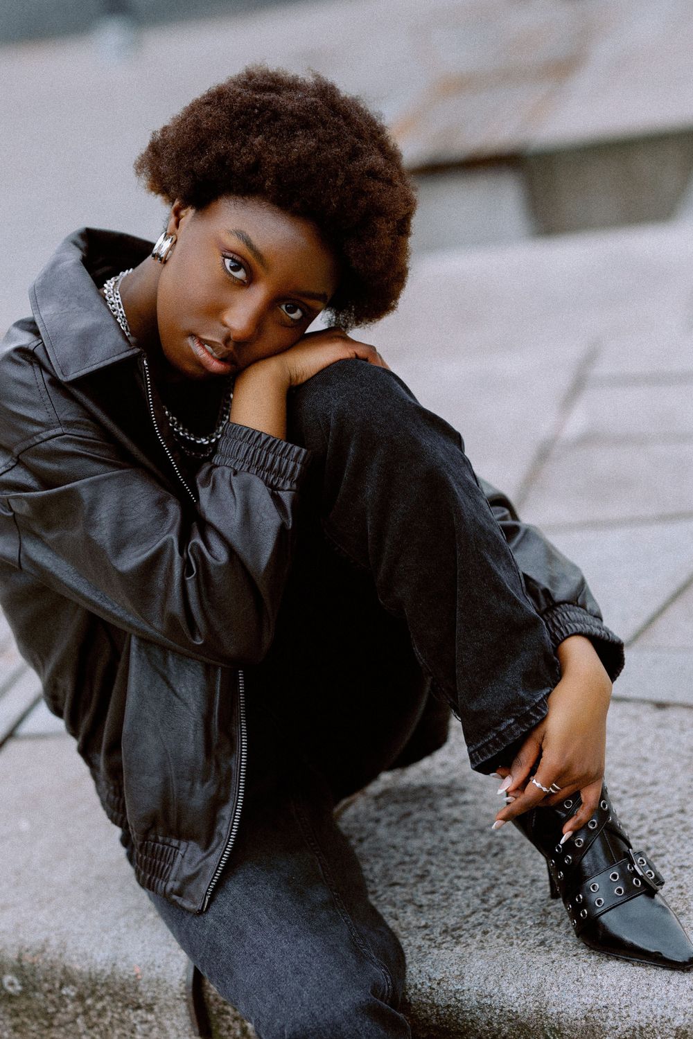 A child in a black leather jacket poses artistically on concrete steps in an urban setting.
