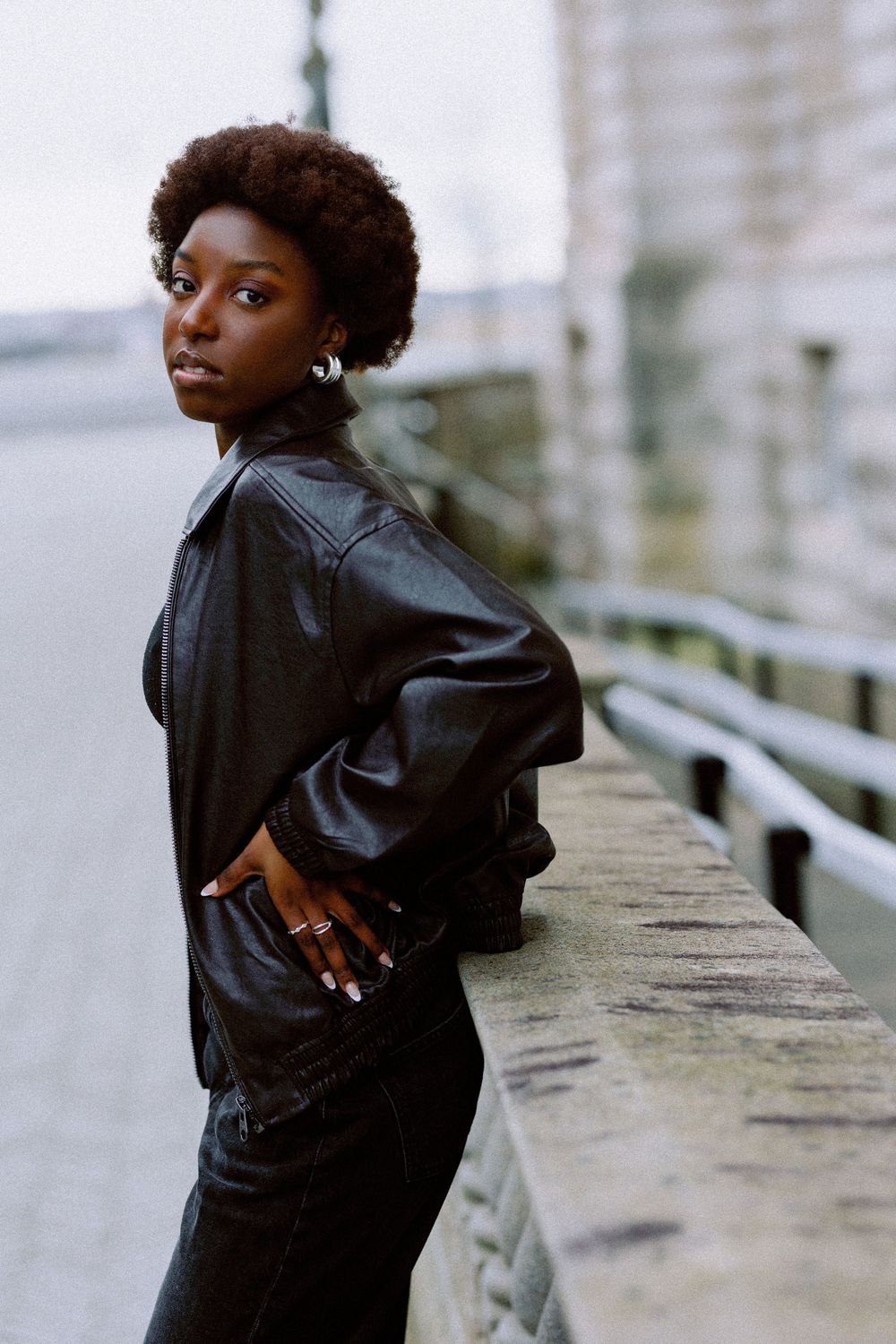 Model in black leather jacket poses against concrete wall in urban setting.