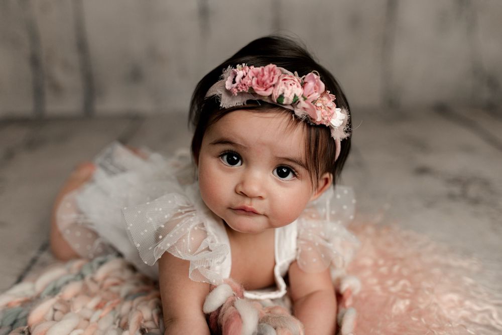 Baby in white lace dress with pink floral headband on textured background.