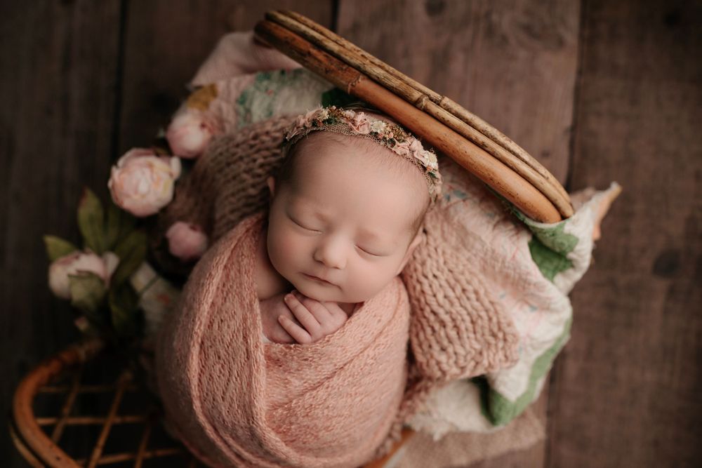 Newborn wrapped in pink swaddle sleeping in rustic basket with flowers.