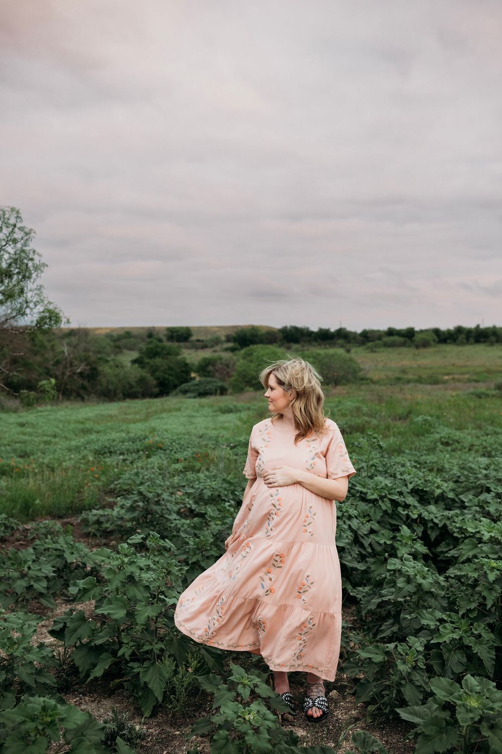 Ethereal outdoor photography in pink dress amid lush green field under cloudy skies.