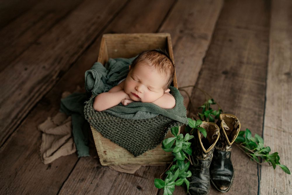 Sleeping newborn in wooden box with vintage boots and greenery decorations.