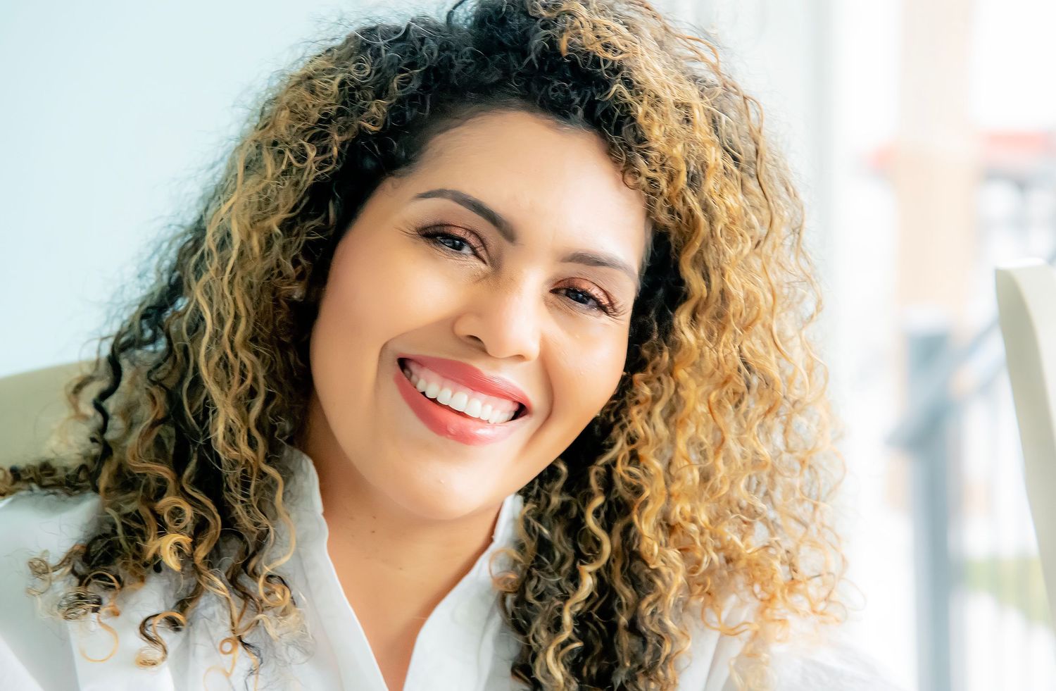 Portrait with natural curly highlighted hair showing bright smile against light background.
