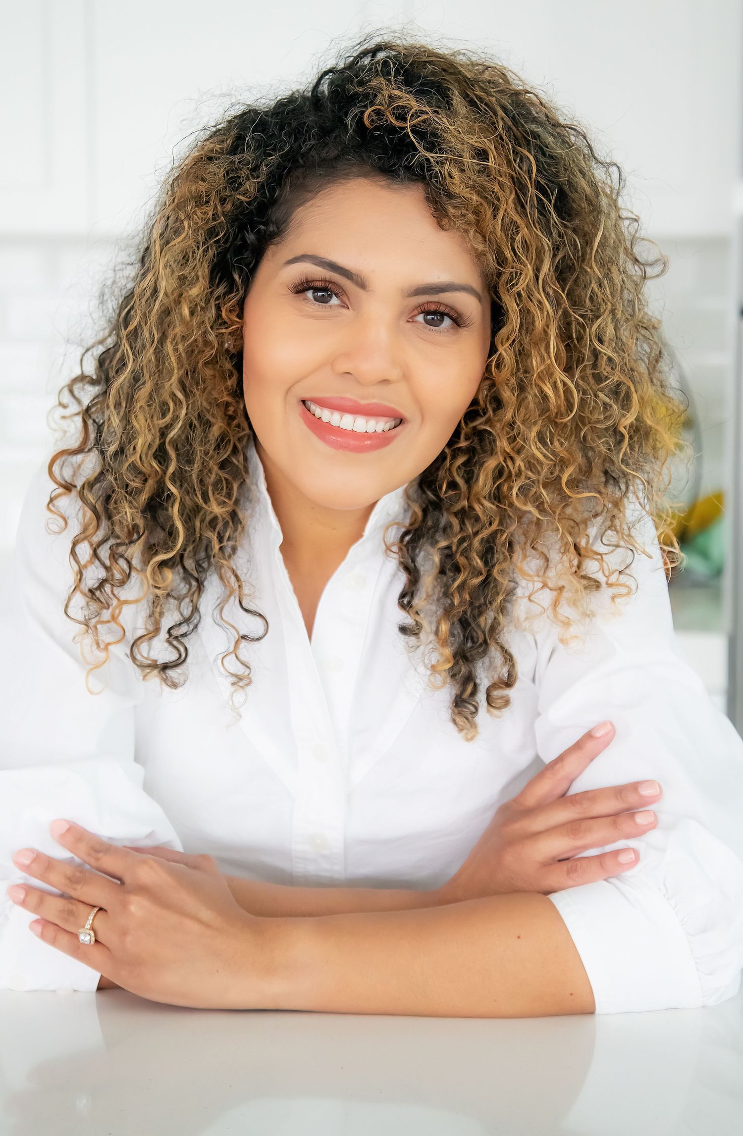 Professional headshot of a person in a white shirt with curly ombre hair smiling at the camera.