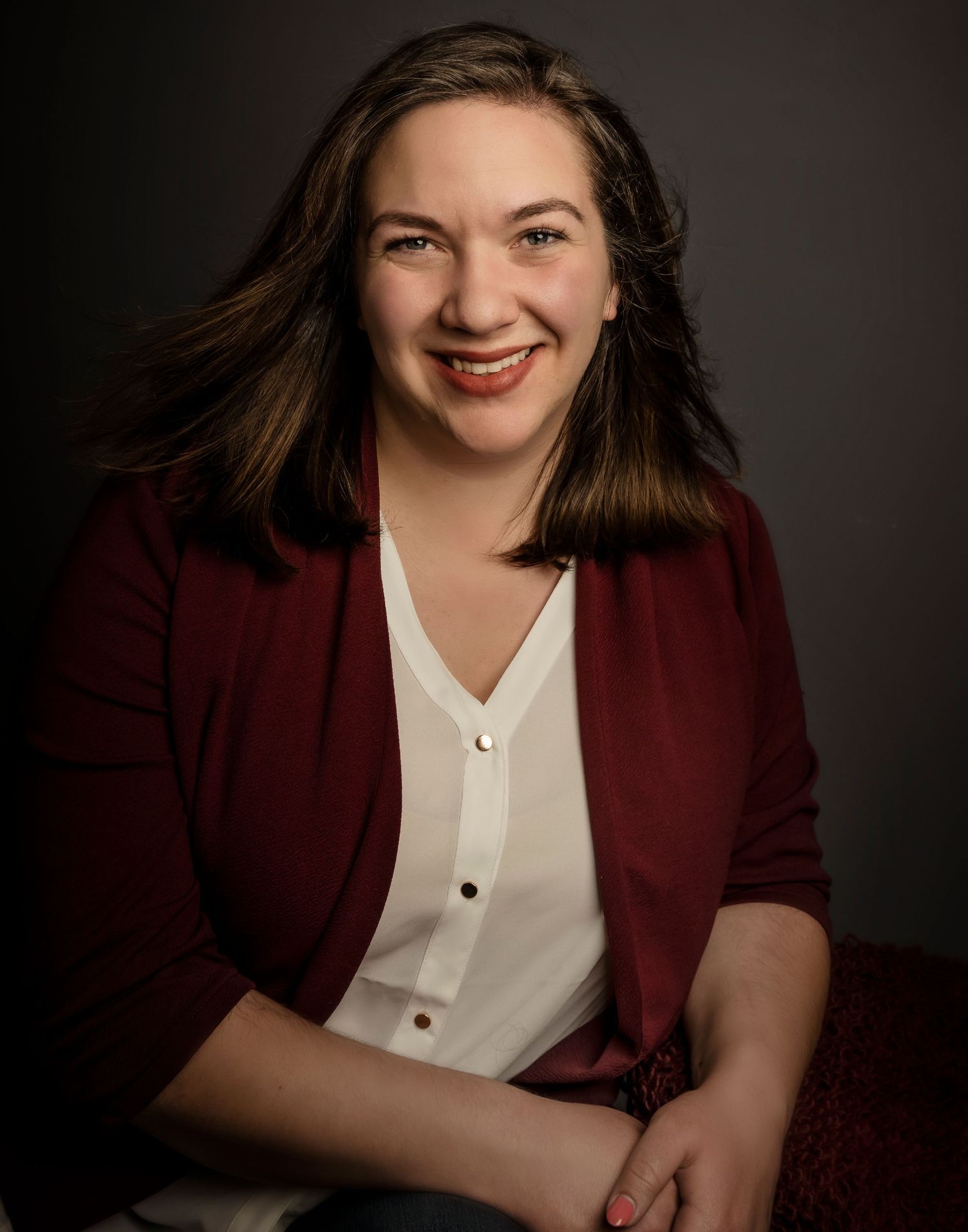 Professional portrait of a person in a burgundy blazer and white blouse against a dark background.