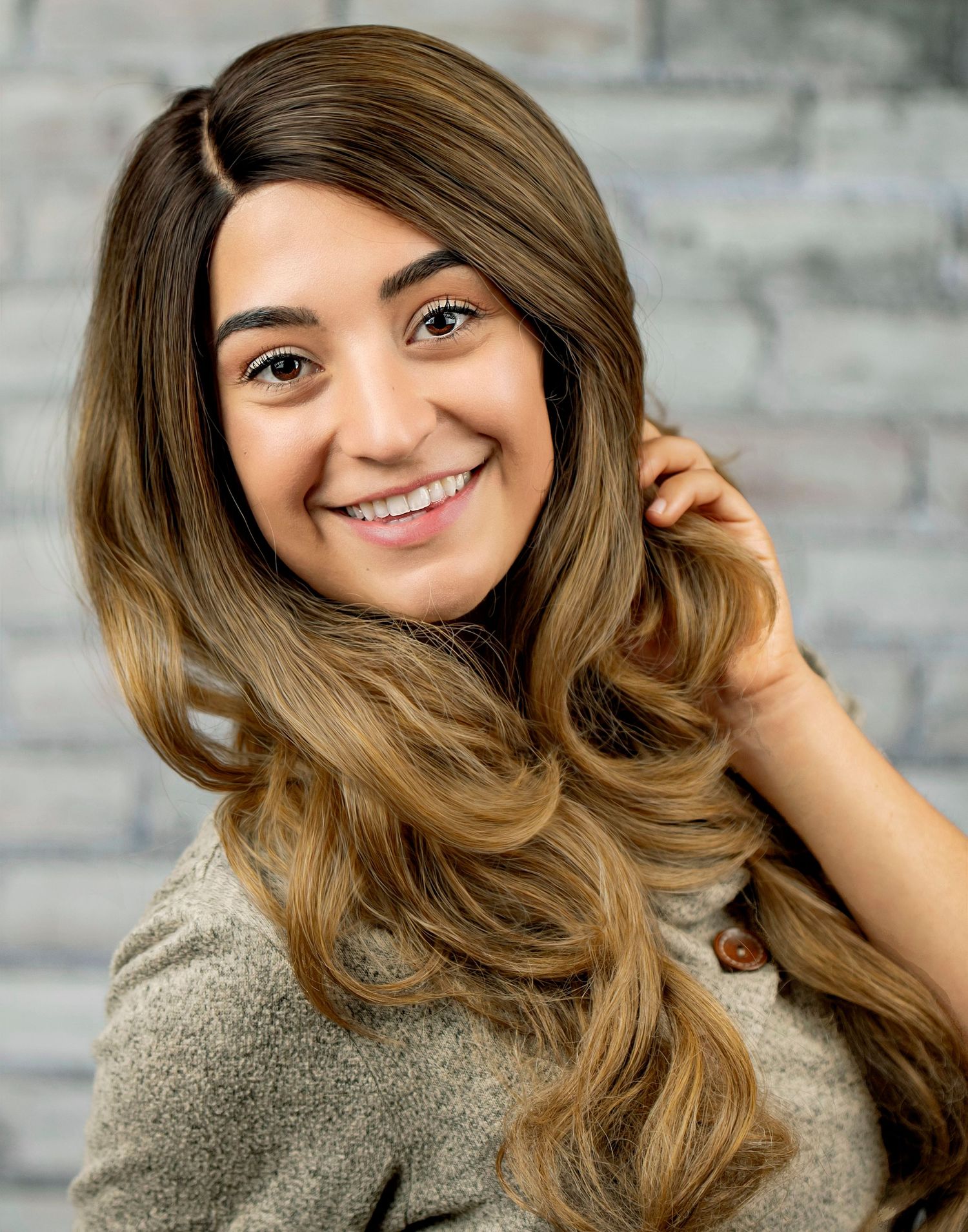 Person with long wavy balayage hair smiling while wearing a gray sweater against a brick wall background.