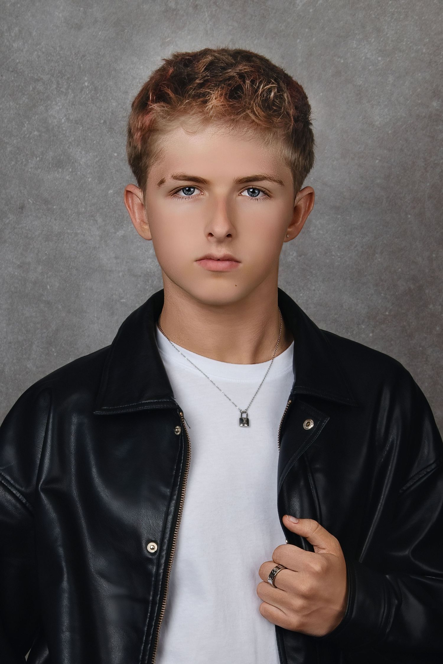 Young man in black leather jacket and white tee with necklace poses confidently against gray studio backdrop.