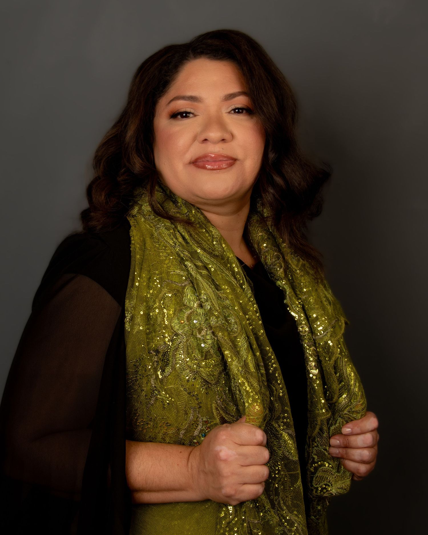 A person wearing a sparkly olive scarf and black top poses against a dark background.