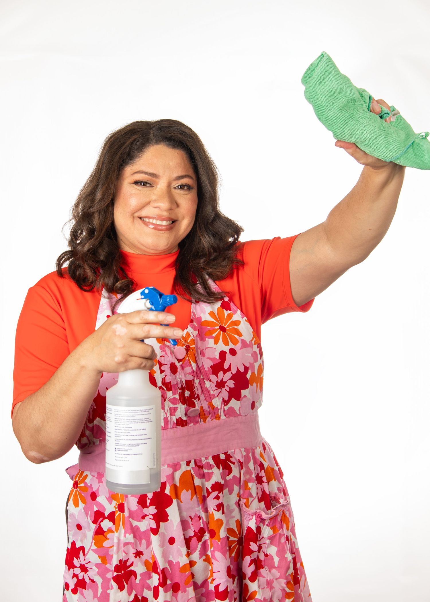 Someone in an orange shirt and floral apron holds cleaning supplies while smiling at the camera.