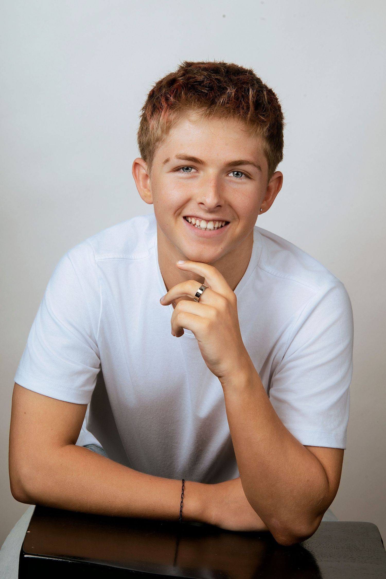 Smiling teenage boy in white t-shirt resting chin on hand at dark table against light gray background.