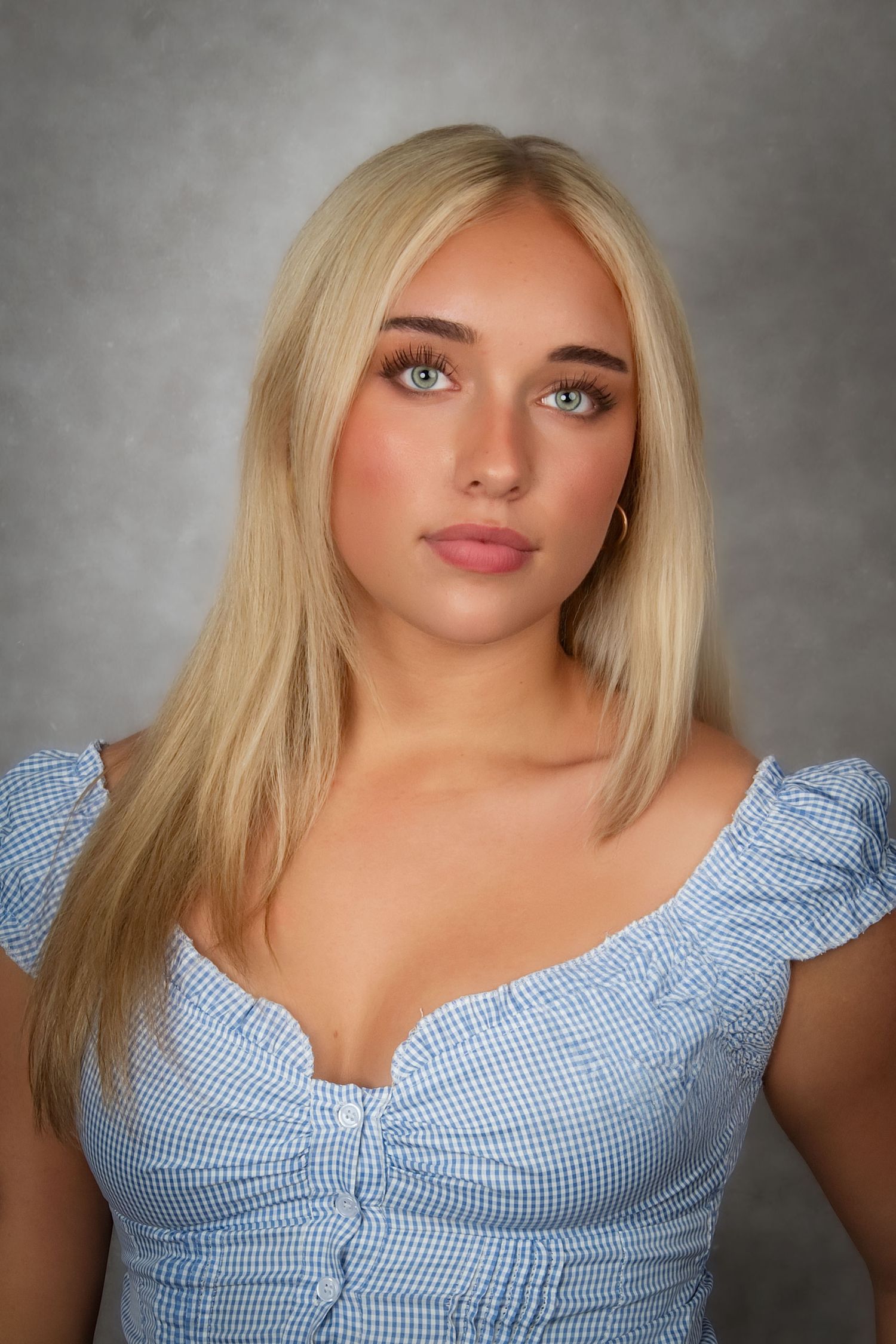 Young blonde woman with blue eyes wearing a blue gingham dress poses for a portrait against a gray background.