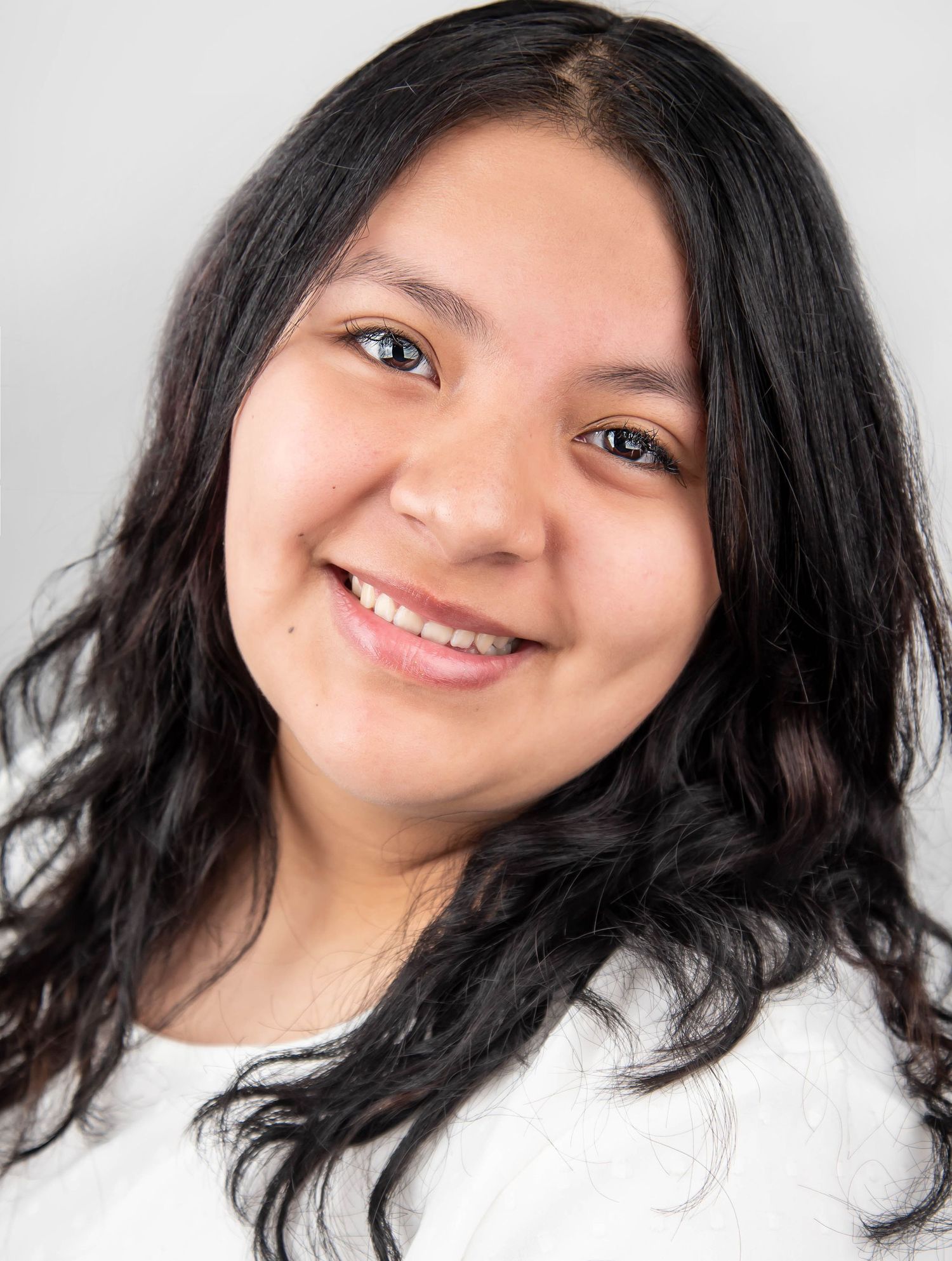 Professional headshot of a person with long dark hair wearing a white top against a light background.