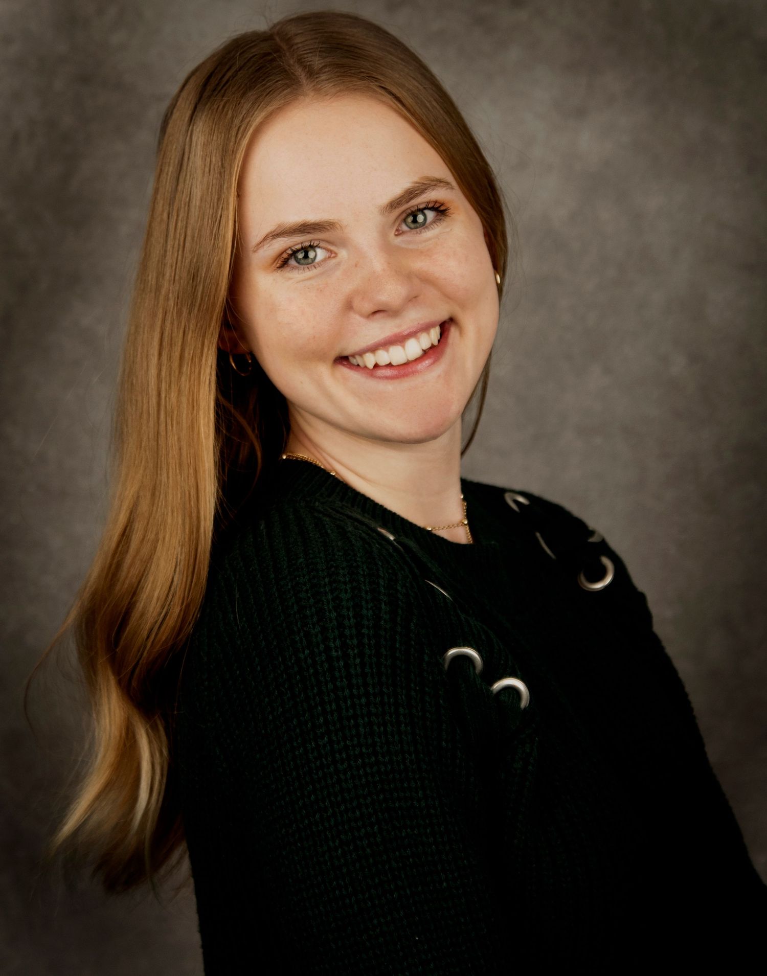 Studio portrait of a person with straight blonde hair wearing a black sweater with grommet details.