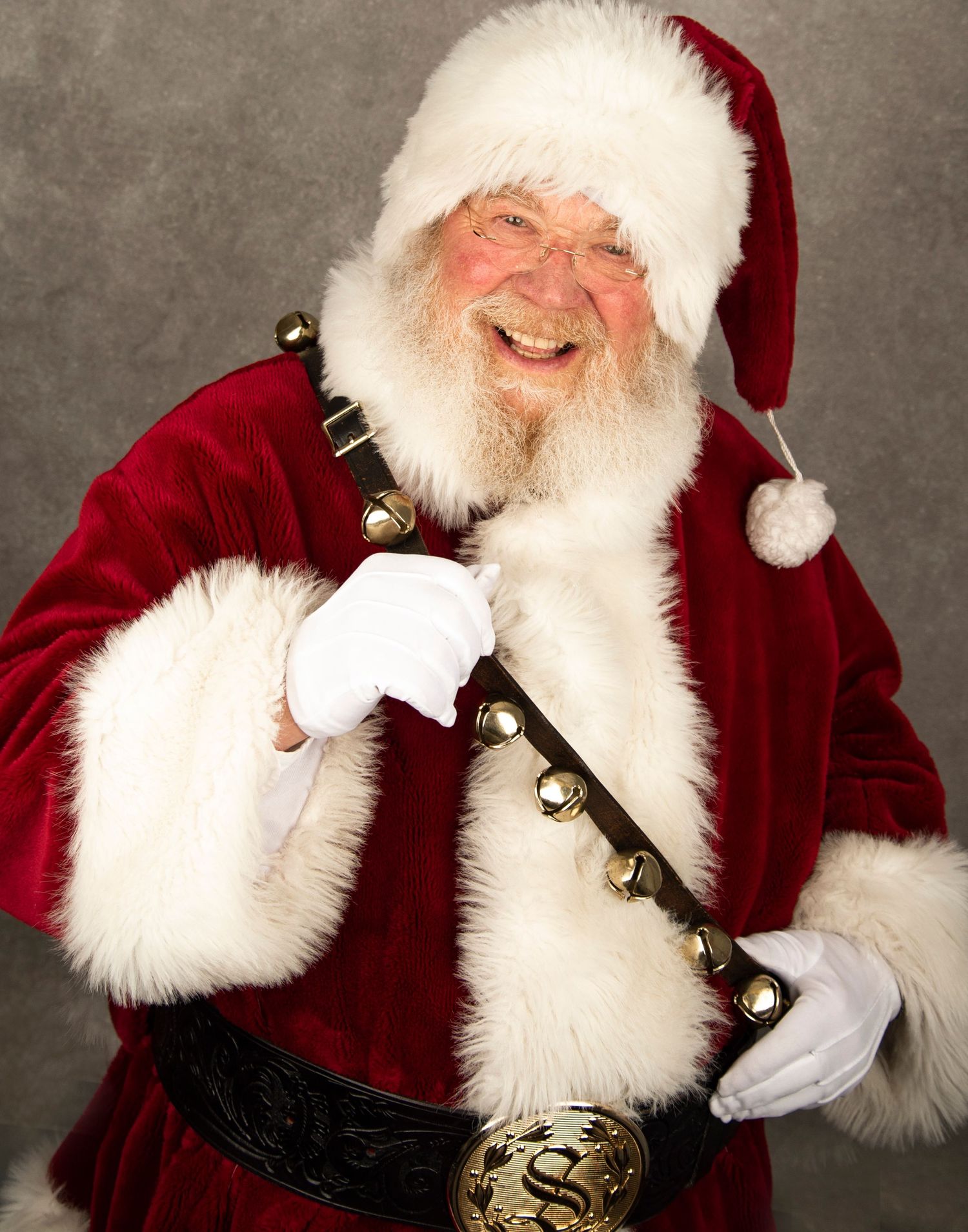 Traditional Santa Claus in red velvet suit trimmed with white fur and jingle bell sash laughs joyfully against gray backdrop.
