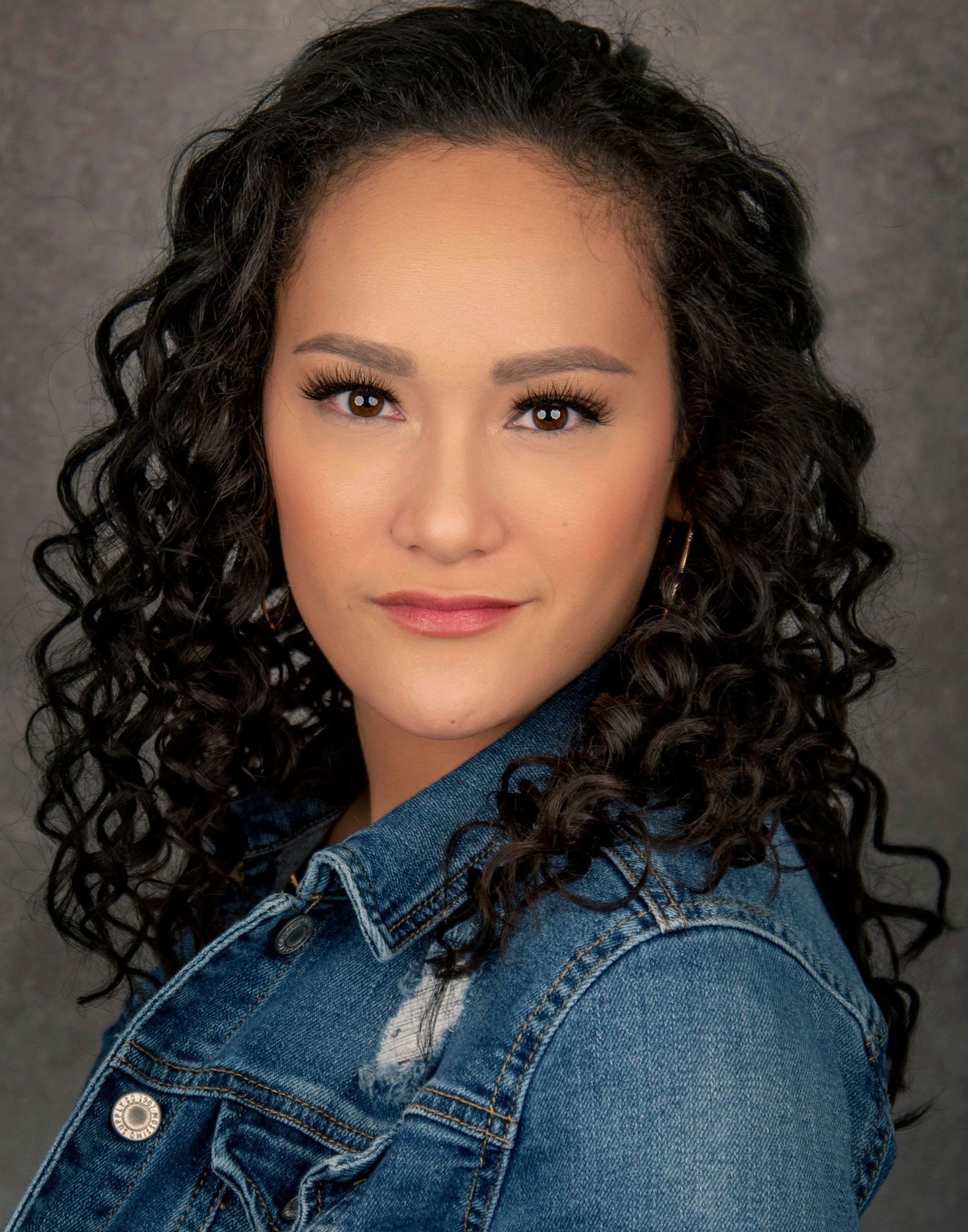 Studio portrait of a person with curly dark hair wearing a denim jacket against a dark backdrop.