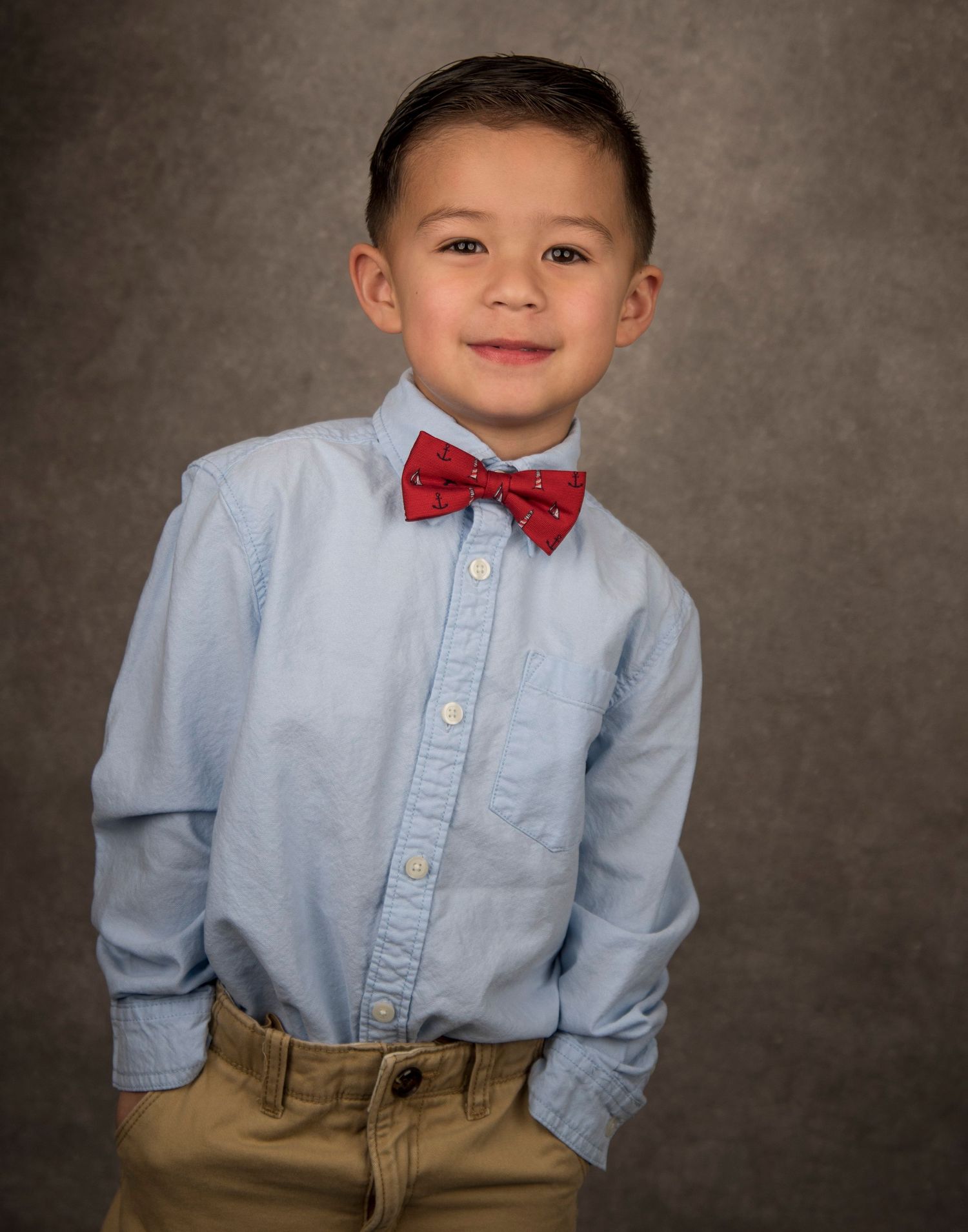 Young child wearing a light blue button-down shirt with a red bowtie and khaki pants poses for a portrait against gray backdrop.