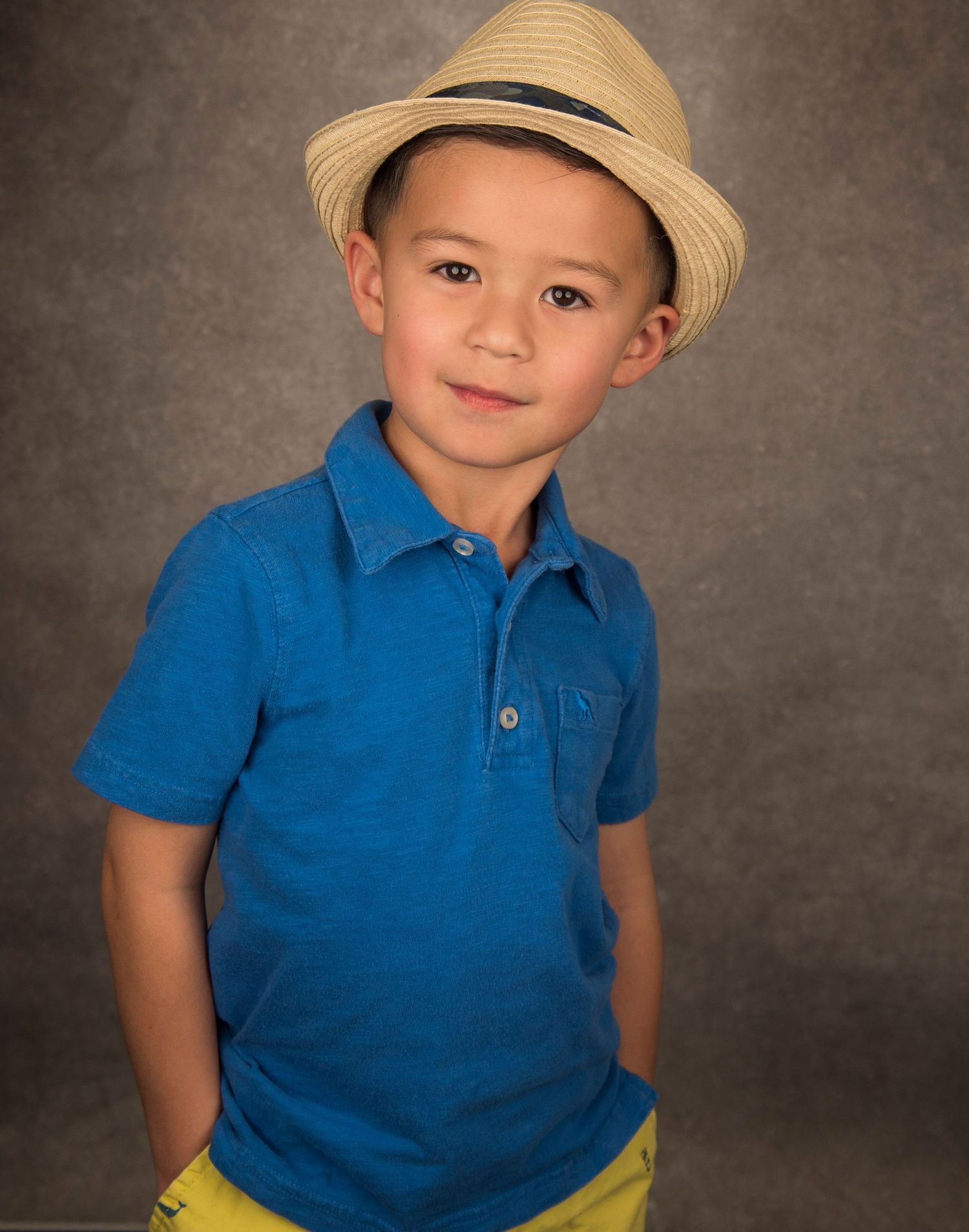 Young child wearing a straw hat and blue polo shirt poses against a textured brown background.