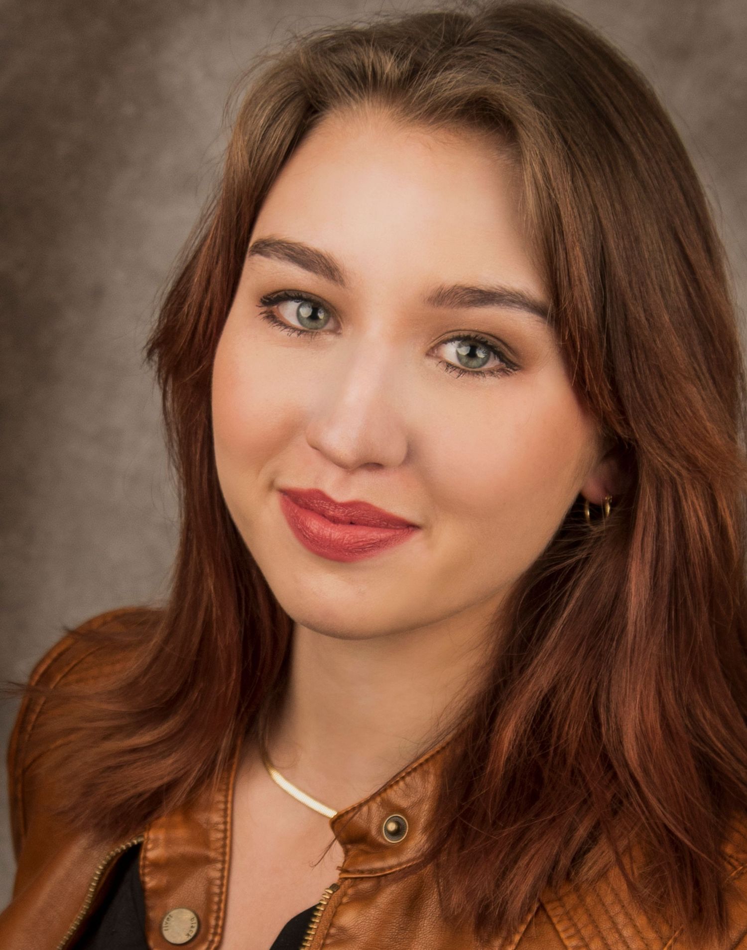 Studio portrait featuring a person in a brown jacket with flowing brown hair and delicate necklace.
