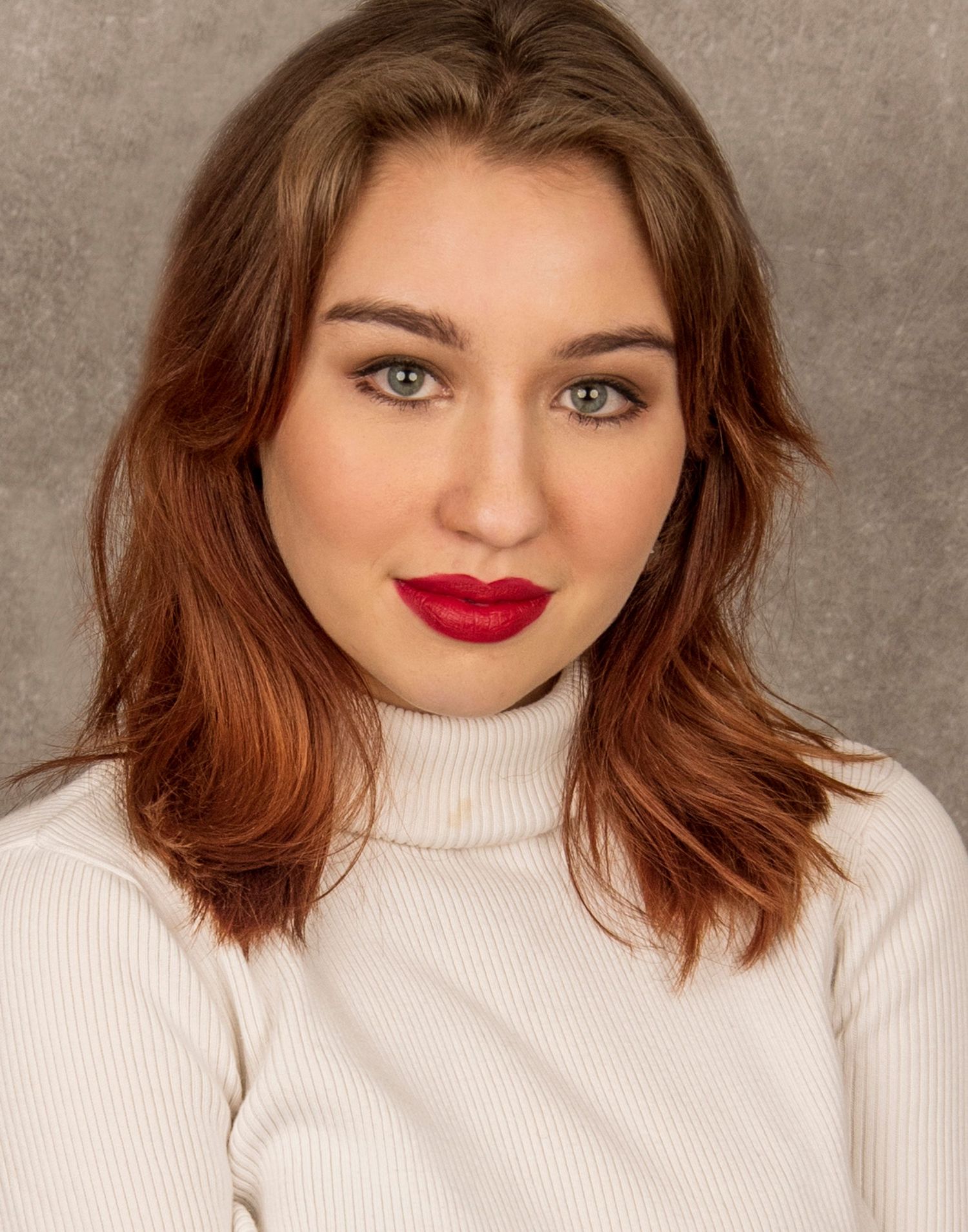 Portrait of a model wearing a white turtleneck sweater, featuring brown shoulder-length hair and bold red lipstick.