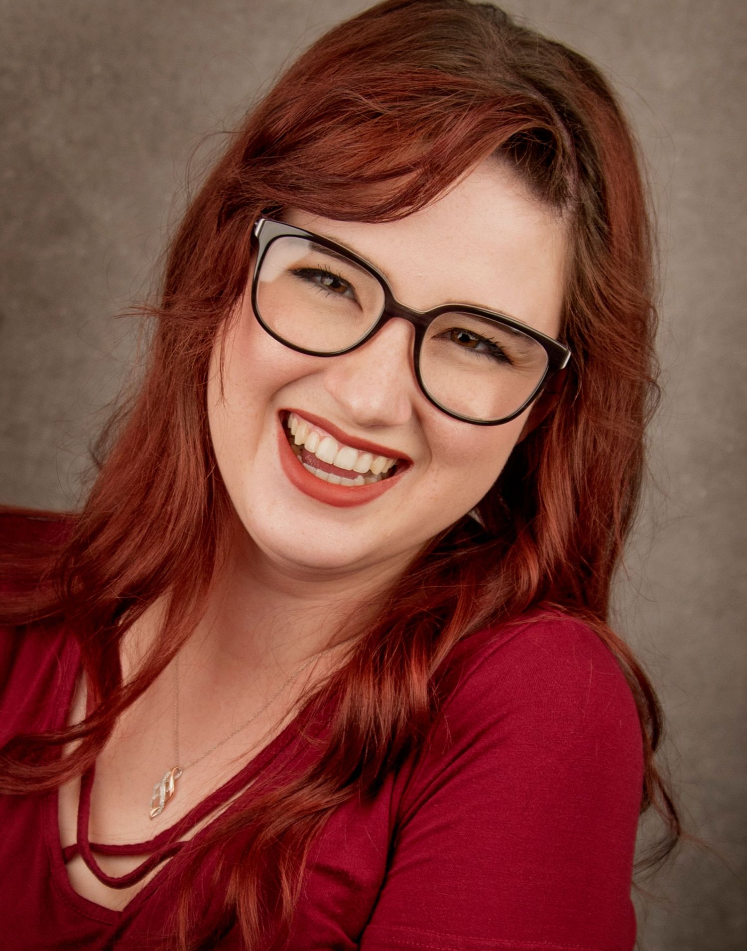 Professional headshot of a person with vibrant red hair wearing glasses and a burgundy top against a neutral background.