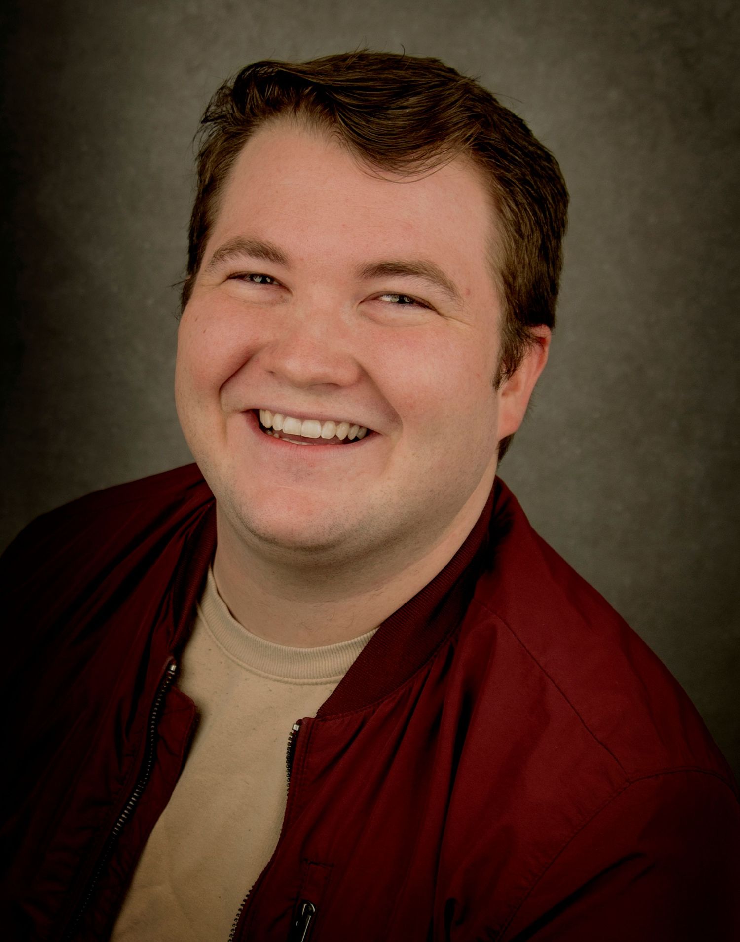 Professional headshot portrait of someone wearing a burgundy jacket and beige shirt against a dark backdrop.