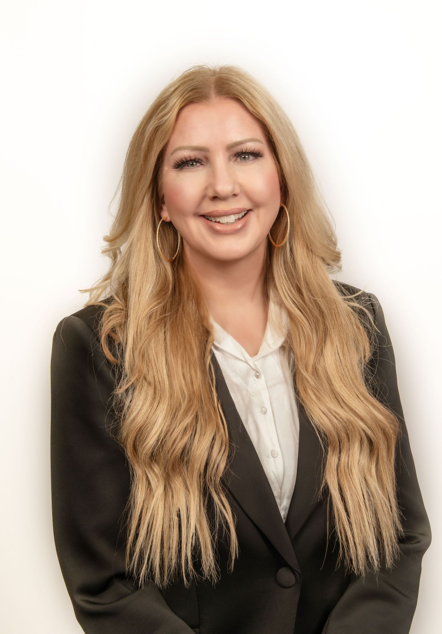 Professional headshot of someone with long blonde hair wearing a black blazer and white blouse against a neutral background.