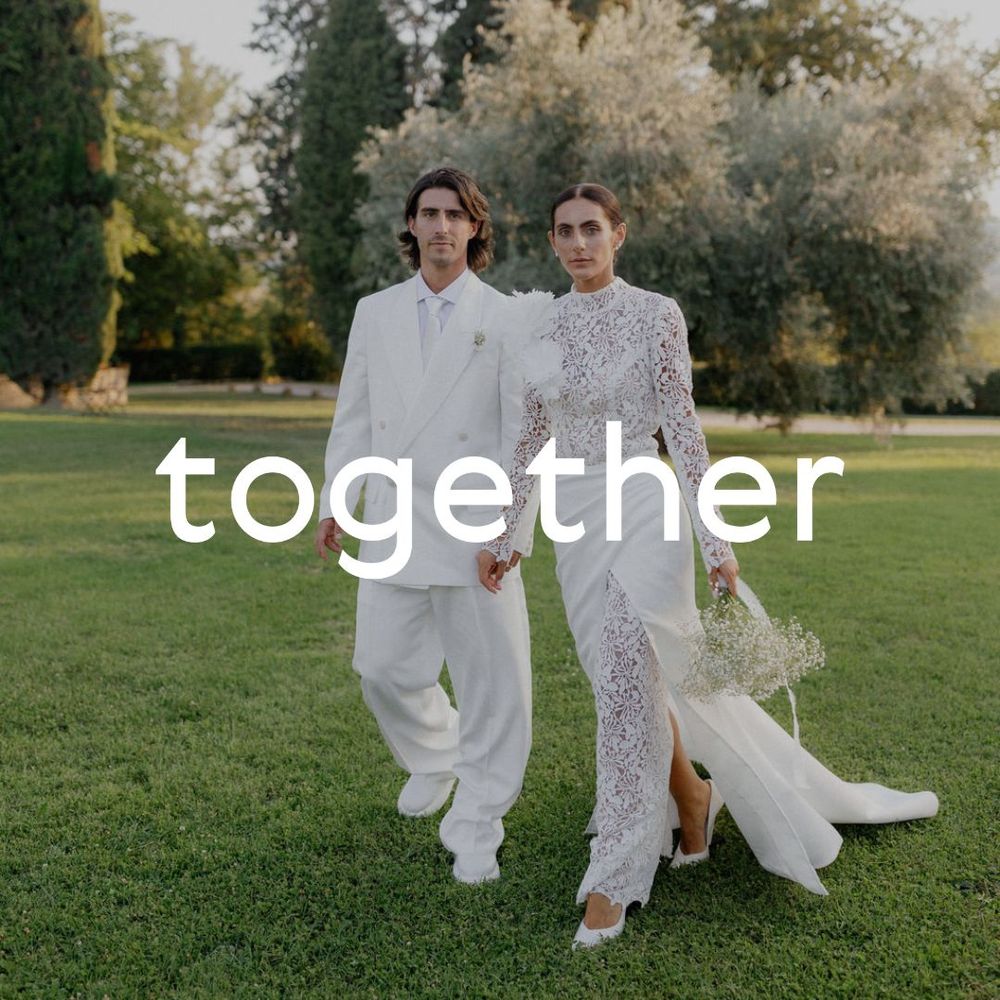 Minimalist wedding photo of couple in white outfits walking together on green lawn at sunset.