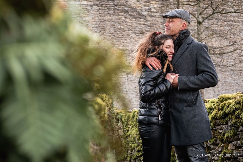 Couple embracing outdoors near a moss-covered stone wall surrounded by lush greenery on a cloudy day.