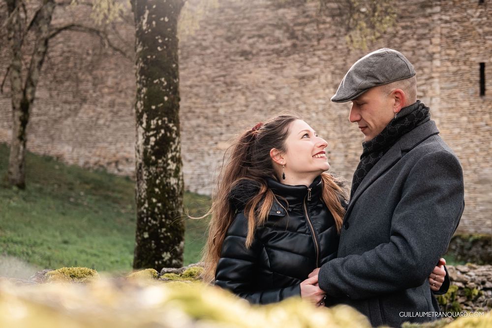 A couple smiles at each other outdoors near a stone wall, surrounded by moss and trees in a romantic setting.