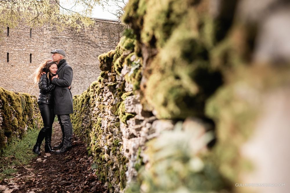 Couple embracing romantically beside a moss-covered stone wall in a lush garden setting.