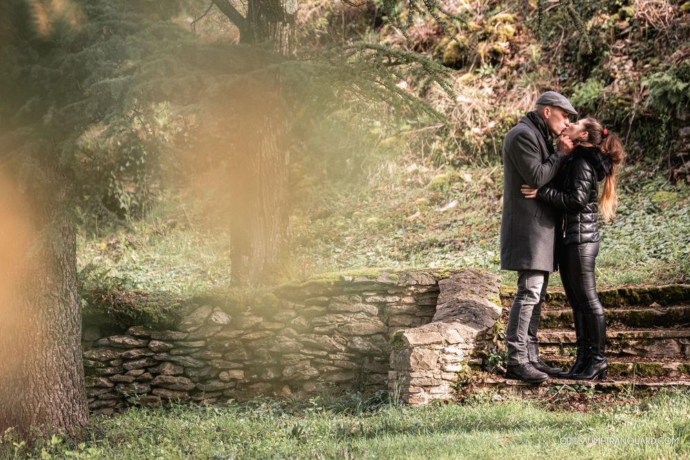Couple sharing a romantic moment in an autumn park beside a rustic stone wall surrounded by lush greenery.