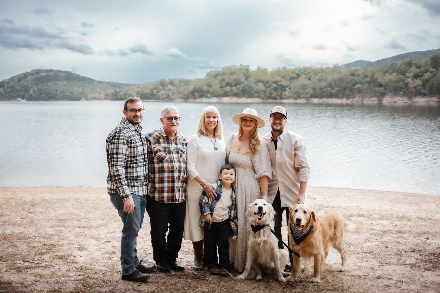 Family and dogs pose together on beach with mountain lake backdrop.